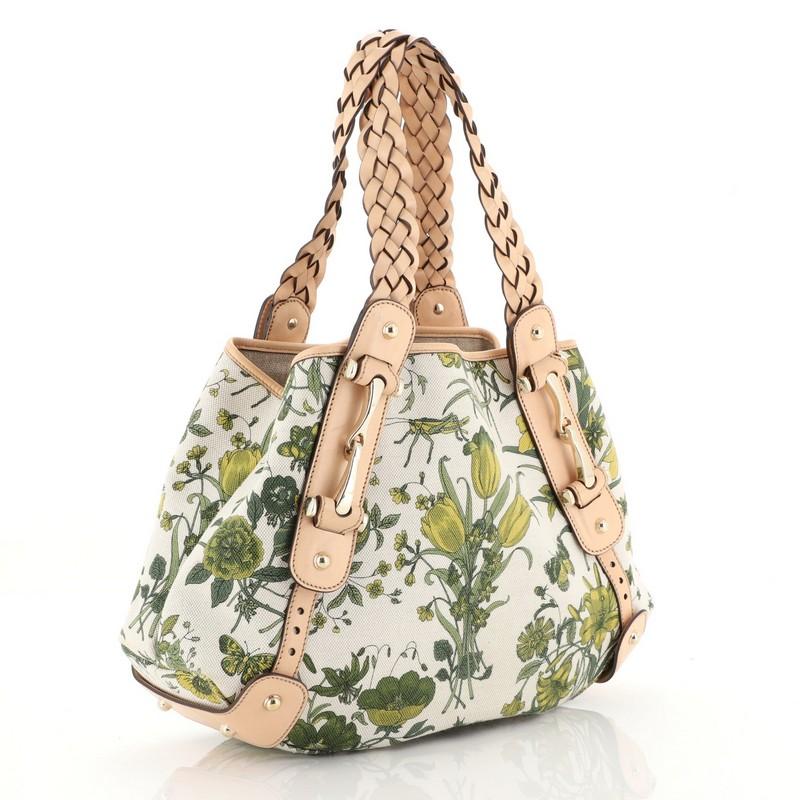 This Gucci Pelham Shoulder Bag Flora Canvas Small, crafted in neutral GG canvas with green floral print, features braided leather straps, signature horsebit detailing, and gold-tone hardware. It opens to a neutral fabric interior with side zip