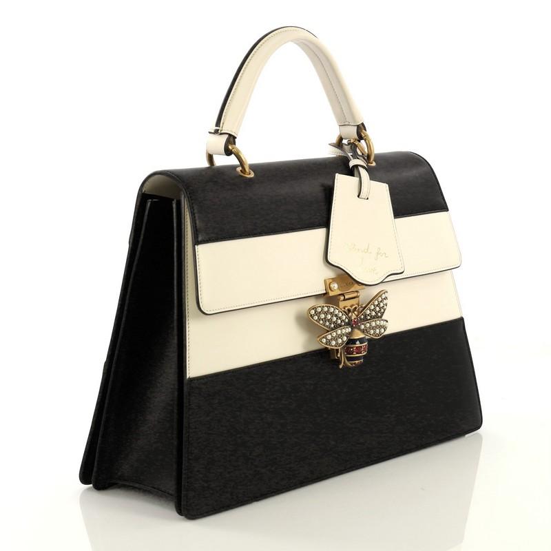 This Gucci Queen Margaret Top Handle Bag Colorblock Leather Large, crafted from black and white leather, features a leather top handle, bejeweled bee on its flap, and aged gold-tone hardware. It opens to a beige microfiber interior divided into two