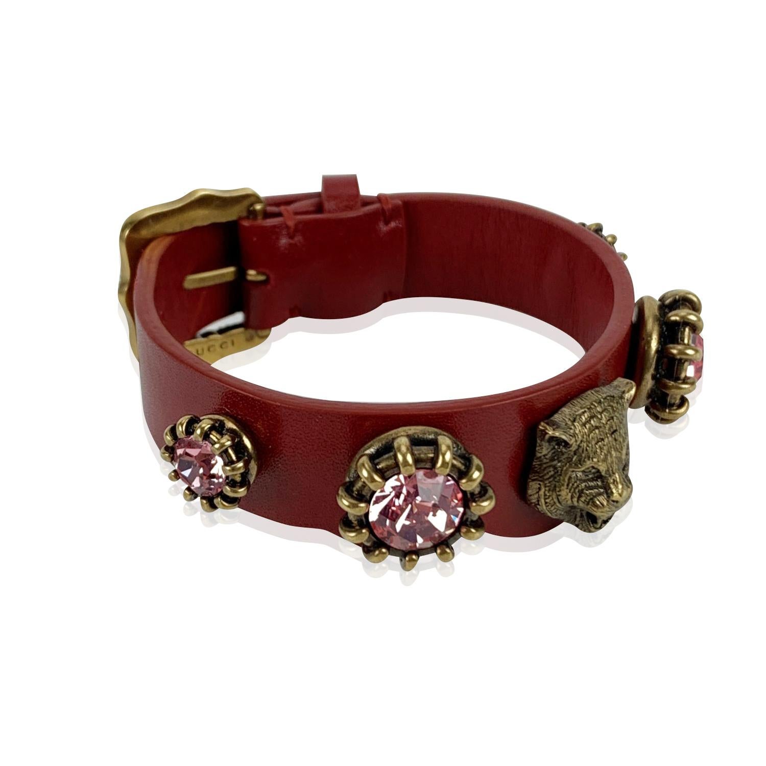 Magnifique bracelet ceinture de Gucci. Confectionné en cuir rouge, il est orné d'une tête de tigre en métal doré vieilli et de détails en cristaux roses. Fermeture à boucle. 3 trous de réglage. Longueur totale (d'un bout à l'autre) : 8,5 pouces -