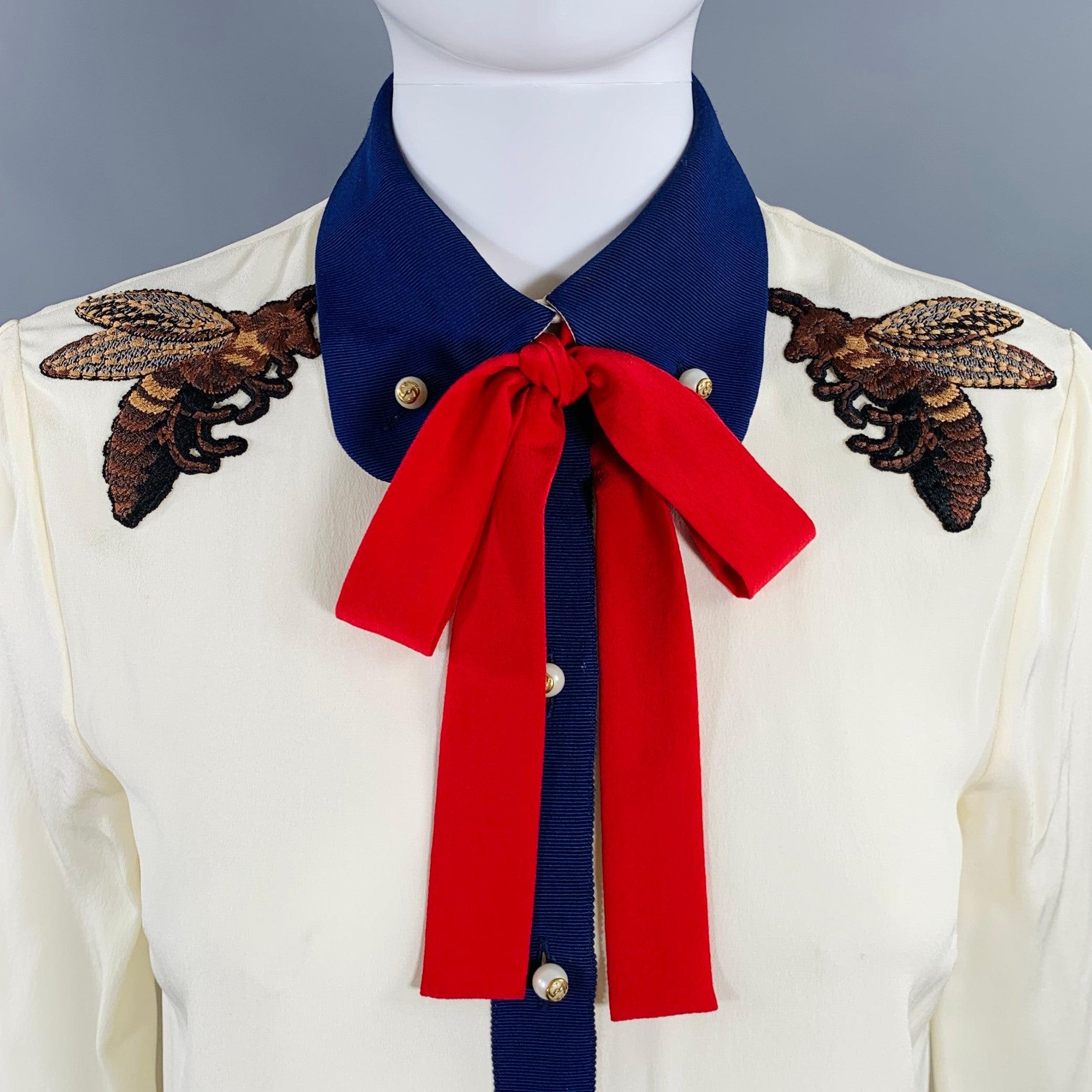 GUCCI blouse
in a cream silk featuring a button down Peter Pan collar, embroidered honeybee patches, red silk pussybow tie, and button closure with faux pearl GUCCI logo buttons. Made in Italy.Very Good Pre-Owned Condition. Minor marks on front and