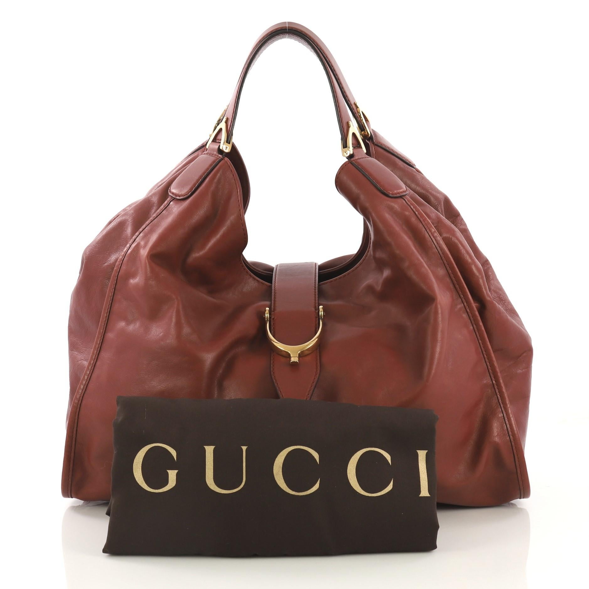 This Gucci Soft Stirrup Tote Leather Large, crafted from dark red leather, features dual flat handles with spur detailing and gold-tone hardware. Its slide flap closure opens to a beige fabric interior with side zip and slip pockets. 

Estimated