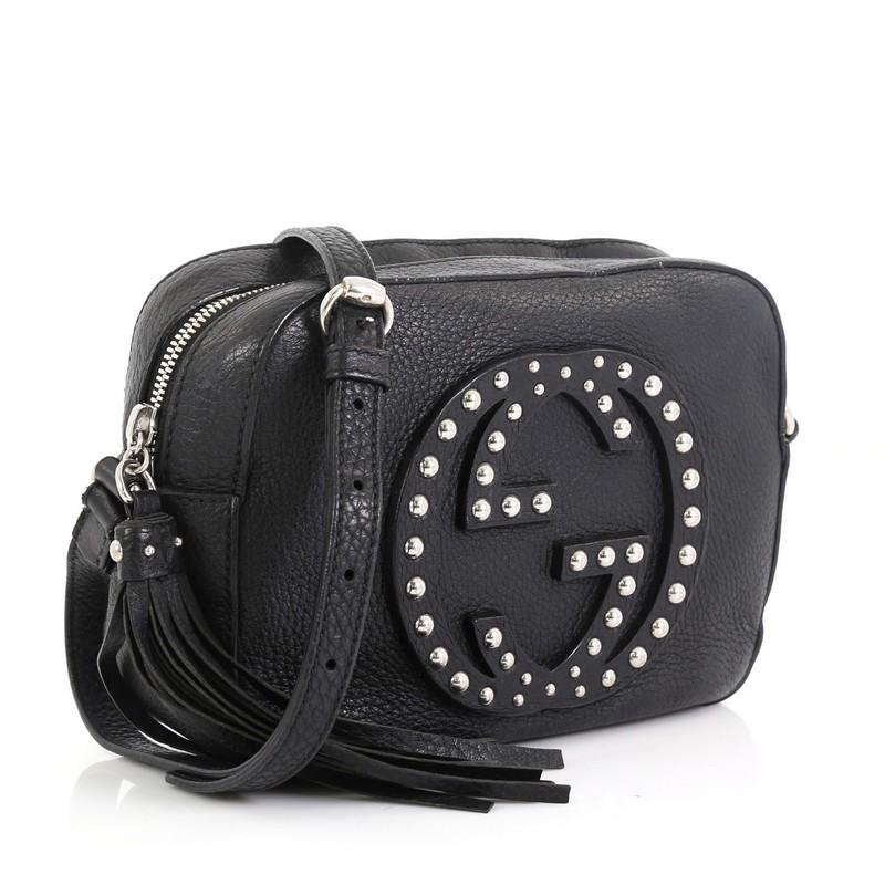 This Gucci Soho Disco Crossbody Bag Studded Leather Small, crafted in black leather, features an adjustable leather strap, tassel zip pull, studded interlocking GG logo on front, and silver-tone hardware. Its zip closure opens to a neutral fabric