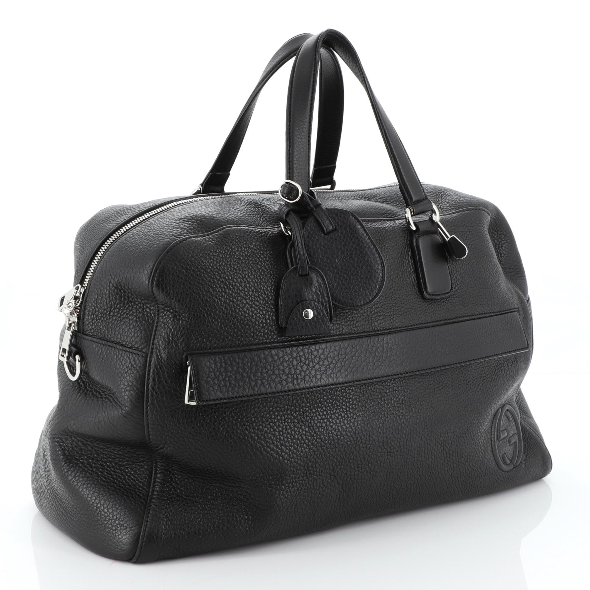 This Gucci Soho Duffle Bag Leather Large, crafted in black leather, features dual flat leather handles, front zip pocket, protective base studs, and silver-tone hardware. Its top zip closure opens to a neutral fabric interior with side zip and slip