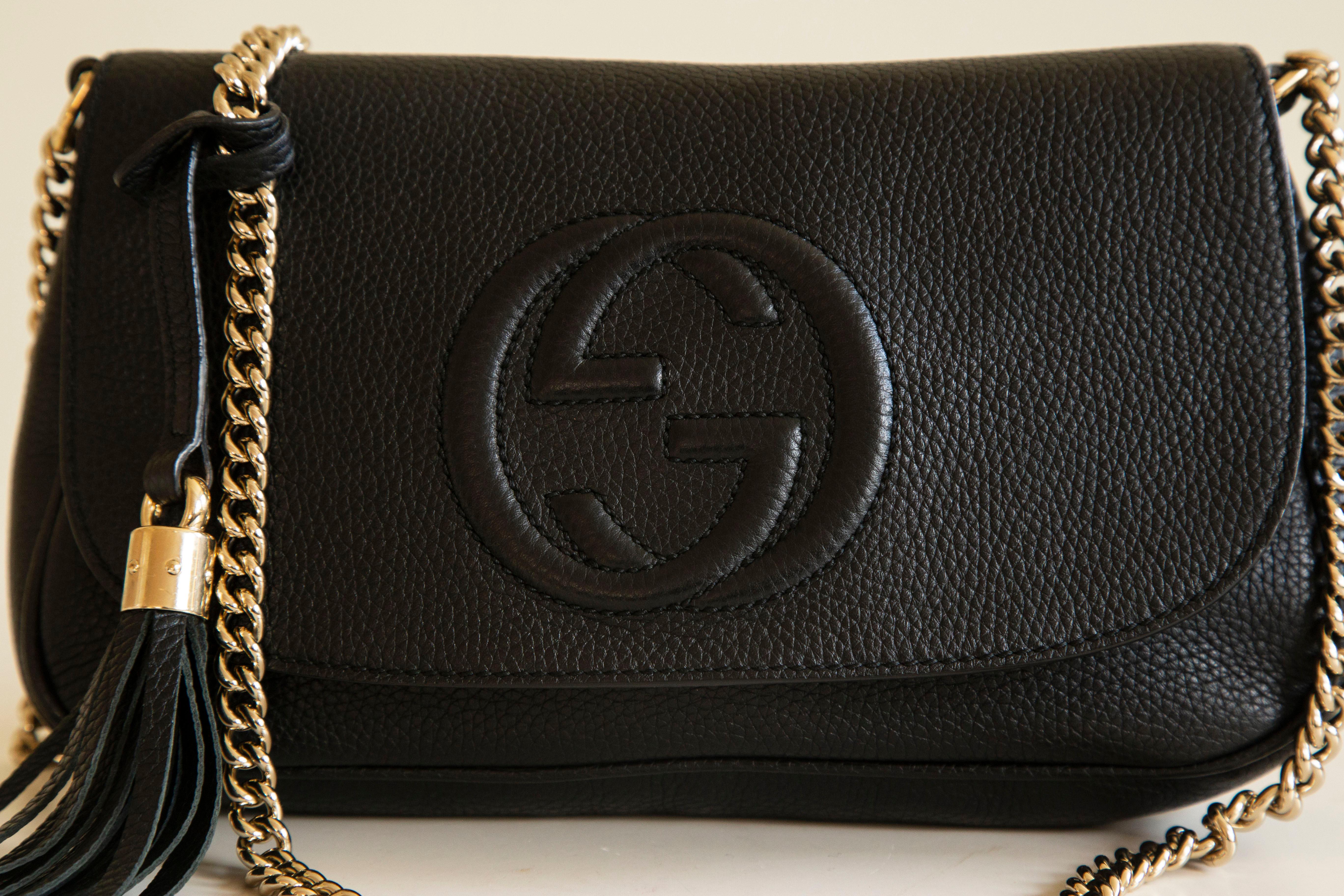 An authentic Gucci Soho crossbody bag in black leather  with chain strap. The bag features a front flap with a magnetic closure. The main compartment is lined with light beige fabric and it contains two side pockets of which one has a zipper. The