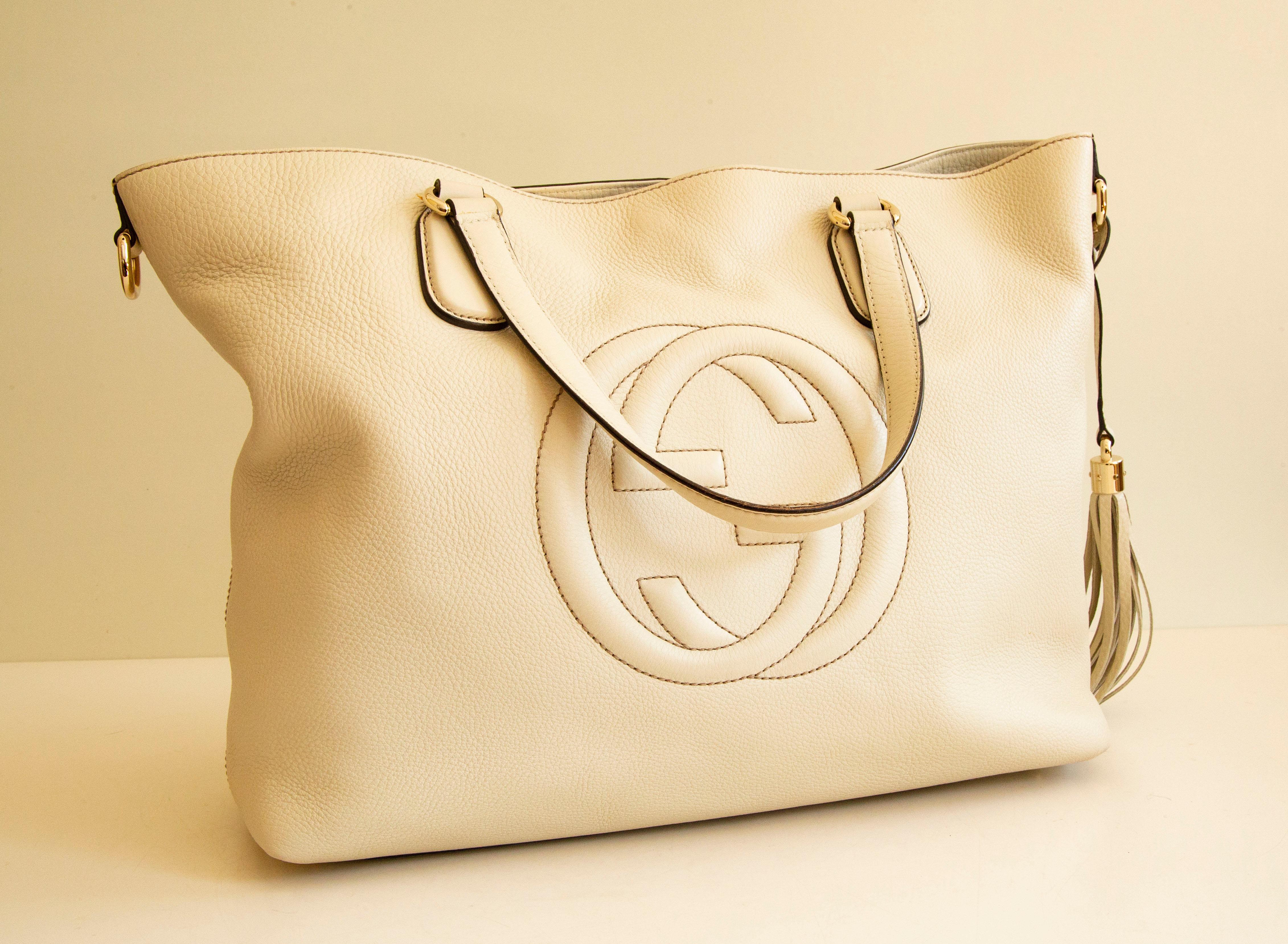 Un sac à main Gucci SOHO en cuir blanc cassé avec des ferrures en or clair. Il est doté d'un pompon en cuir amovible. L'intérieur est doublé de toile beige et, à côté du compartiment principal, il comporte trois poches latérales, dont l'une est