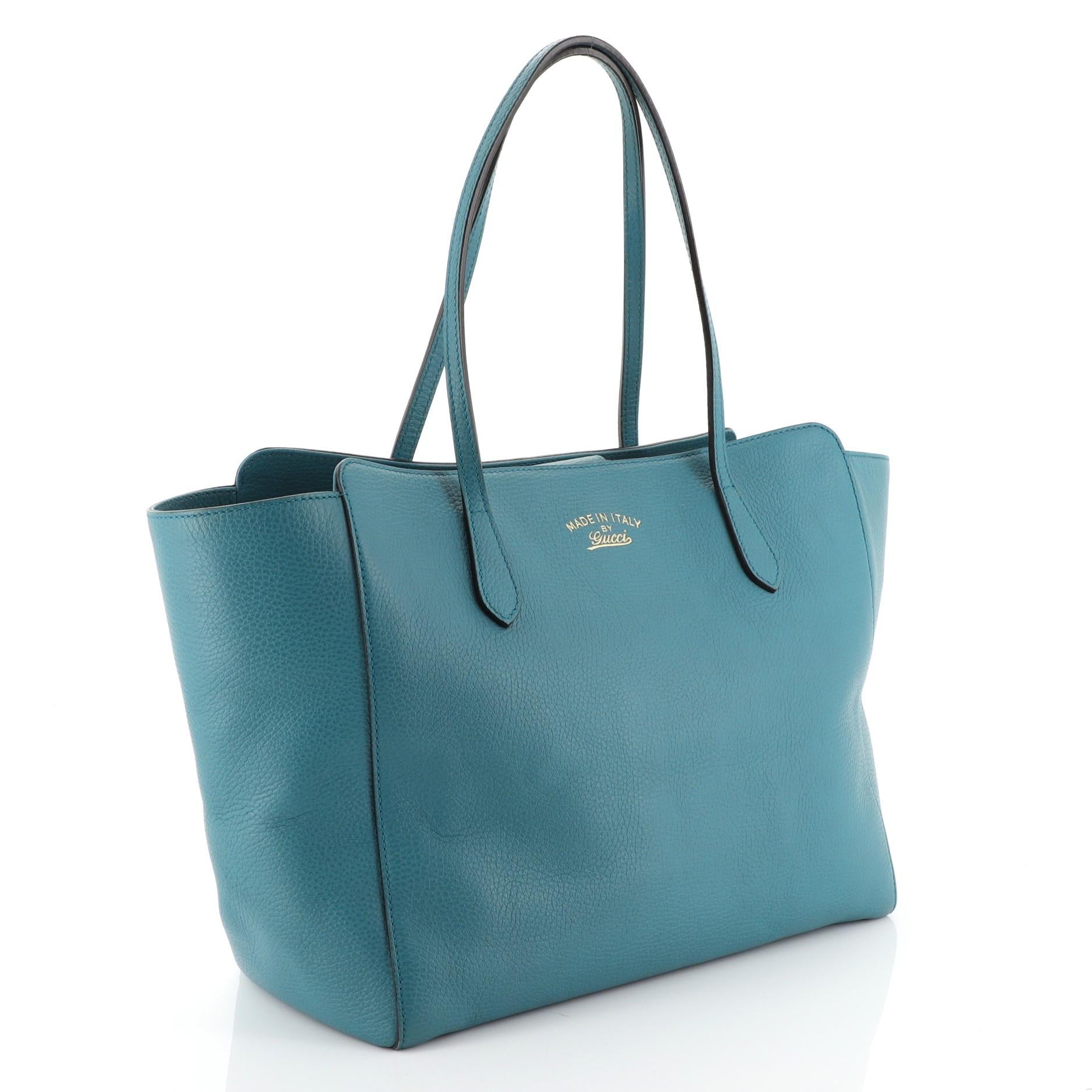 This Gucci Swing Tote Leather Medium, crafted in blue leather, features dual slim handles, Gucci stamped logo at the front, expanded wing silhouette, and gold-tone hardware. Its hidden magnetic snap closure opens to a neutral canvas interior with