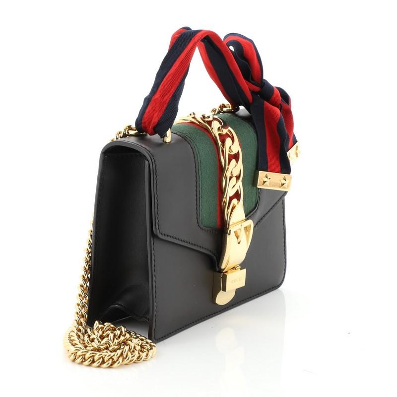 This Gucci Sylvie Chain Shoulder Bag Leather Mini, crafted from black leather, features long chain shoulder strap, grosgrain Web bow, nylon Web detail with curb chain, and gold-tone hardware. Its buckle closure opens to a neutral microfiber interior