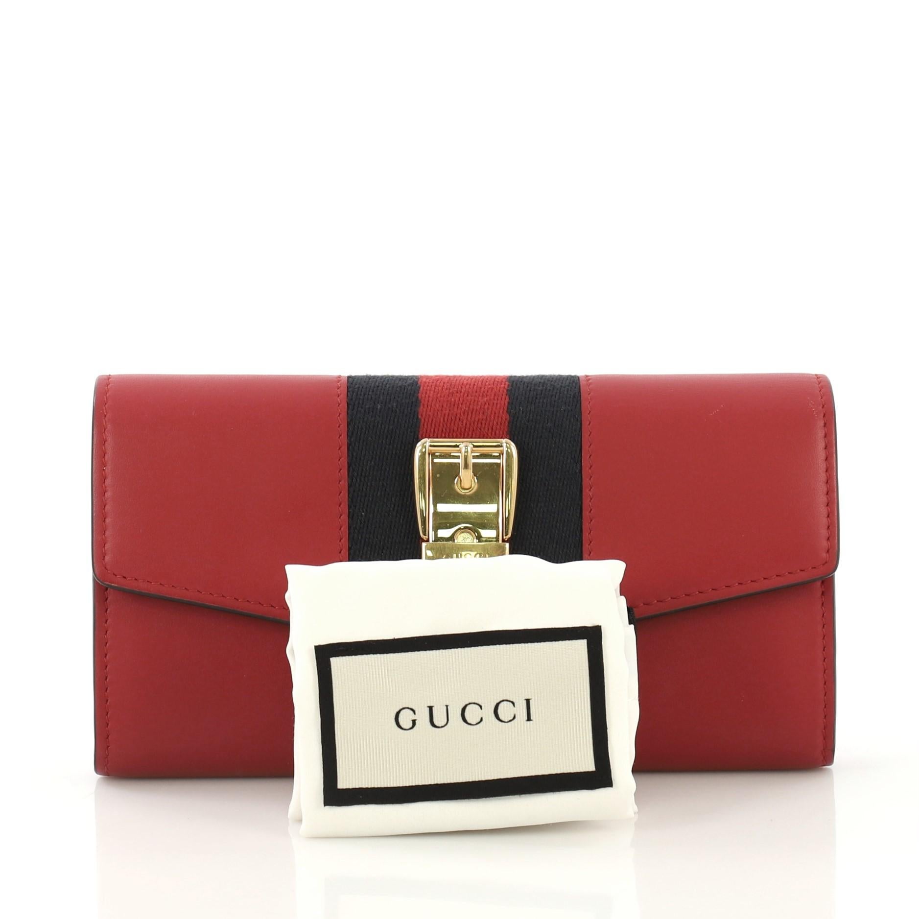 This Gucci Sylvie Continental Wallet Leather, crafted from red leather, features a nylon web detail with buckle accents and gold-tone hardware. It opens to a red leather and black fabric interior with multiple card slots and center zip compartment.