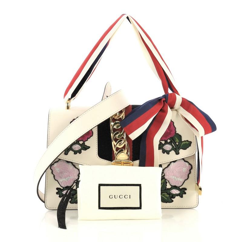 This Gucci Sylvie Shoulder Bag Embroidered Leather Small, crafted in white embroidered leather, features a leather shoulder strap, nylon web detail with curb chain, embroidered flower appliques, and gold-tone hardware. Its buckle closure opens to a