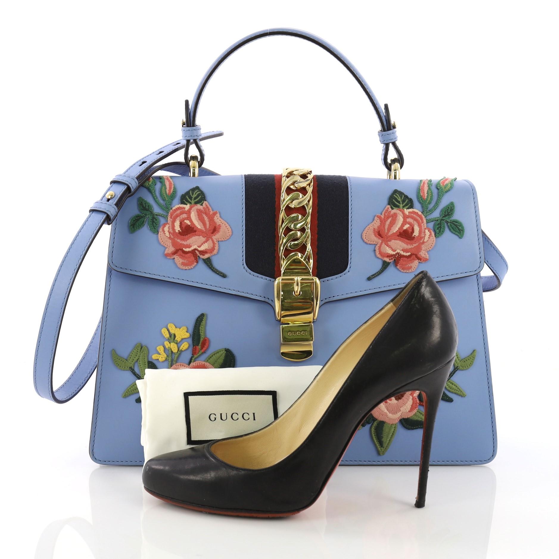 This Gucci Sylvie Top Handle Bag Embroidered Leather Medium, crafted in blue embroidered leather, features a leather shoulder strap, nylon web detail with curb chain, embroidered flower appliques, and gold-tone hardware. Its buckle closure opens to