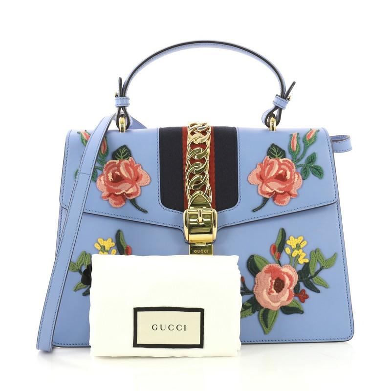 This Gucci Sylvie Top Handle Bag Embroidered Leather Medium, crafted in blue embroidered leather, features a leather top handle, nylon web detail with curb chain, embroidered flowers, and gold-tone hardware. Its buckle closure opens to a brown