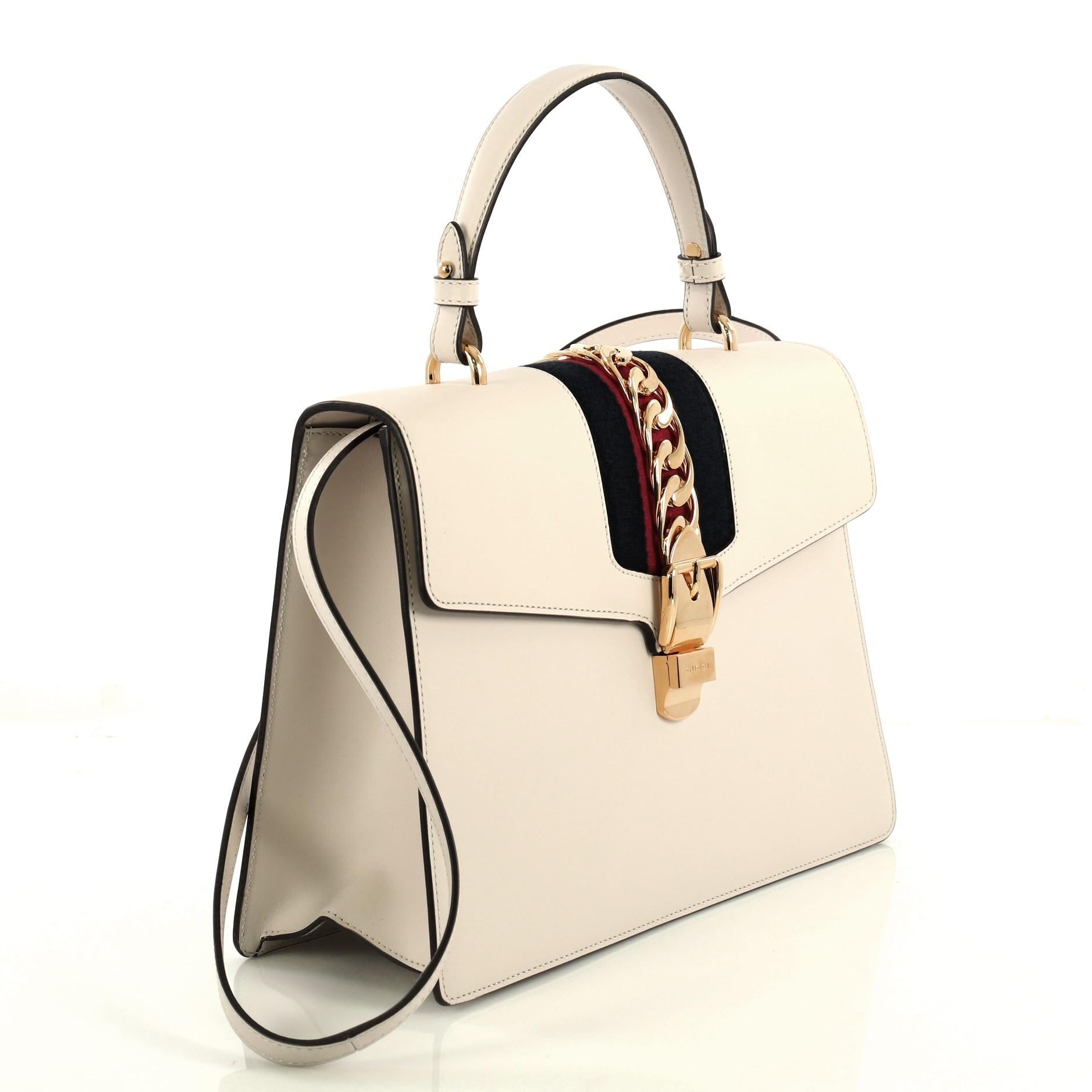 This Gucci Sylvie Top Handle Bag Leather Medium, crafted from white leather, features a single looped leather handle, nylon web detail with curb chain, and gold-tone hardware. Its buckle closure opens to a neutral microfiber interior with side zip