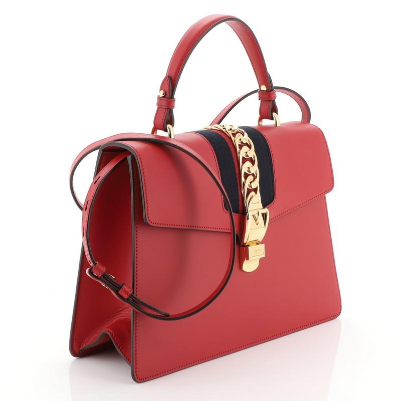 This Gucci Sylvie Top Handle Bag Leather Medium, crafted from red leather, features a single looped leather handle, nylon web detail with curb chain, and gold-tone hardware. Its buckle closure opens to a neutral microfiber interior with side zip and