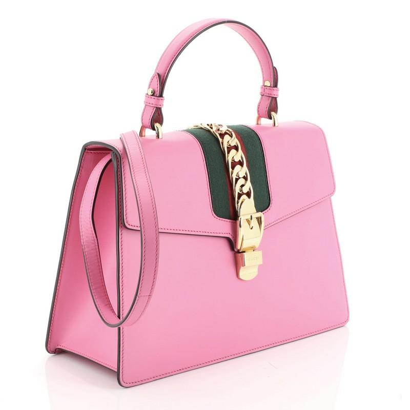 This Gucci Sylvie Top Handle Bag Leather Medium, crafted in pink leather, features single top leather handle, long leather strap, and gold-tone hardware. Its web detail with metal chain and buckle closure opens to a neutral microfiber interior with