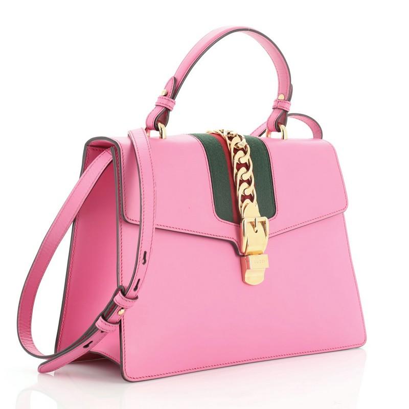 This Gucci Sylvie Top Handle Bag Leather Medium, crafted from pink leather, features a single looped leather handle, nylon web detail with curb chain, and gold-tone hardware. Its buckle closure opens to a neutral microfiber interior with side zip