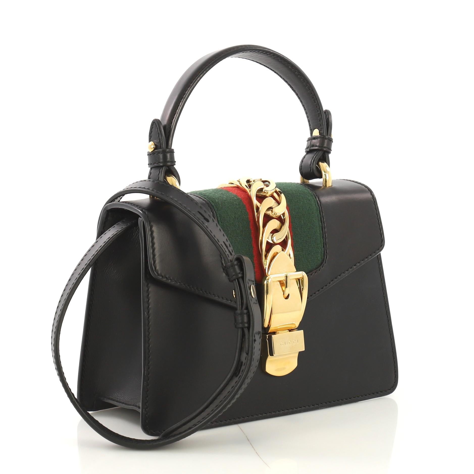 This Gucci Sylvie Top Handle Bag Leather Mini, crafted in black leather, features a single looped leather handle, nylon web detail with curb chain, and gold-tone hardware. Its buckle closure opens to a beige microfiber interior with slip pocket.