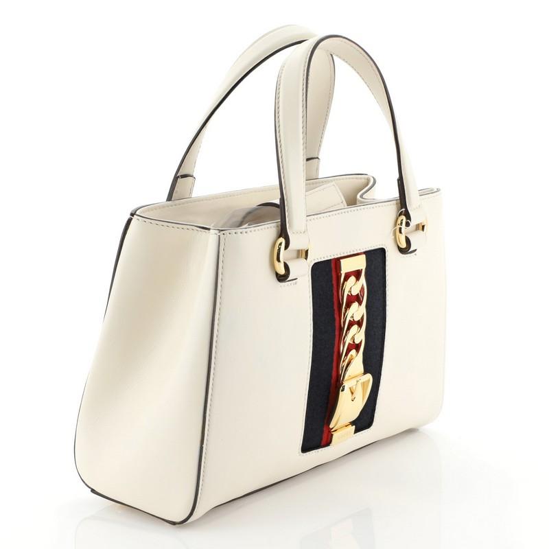This Gucci Sylvie Top Handle Tote Leather Medium, crafted from white leather, features dual leather handles, nylon web detail with curb chain, and gold-tone hardware. Its magnetic snap closure opens to a neutral microfiber interior with two open
