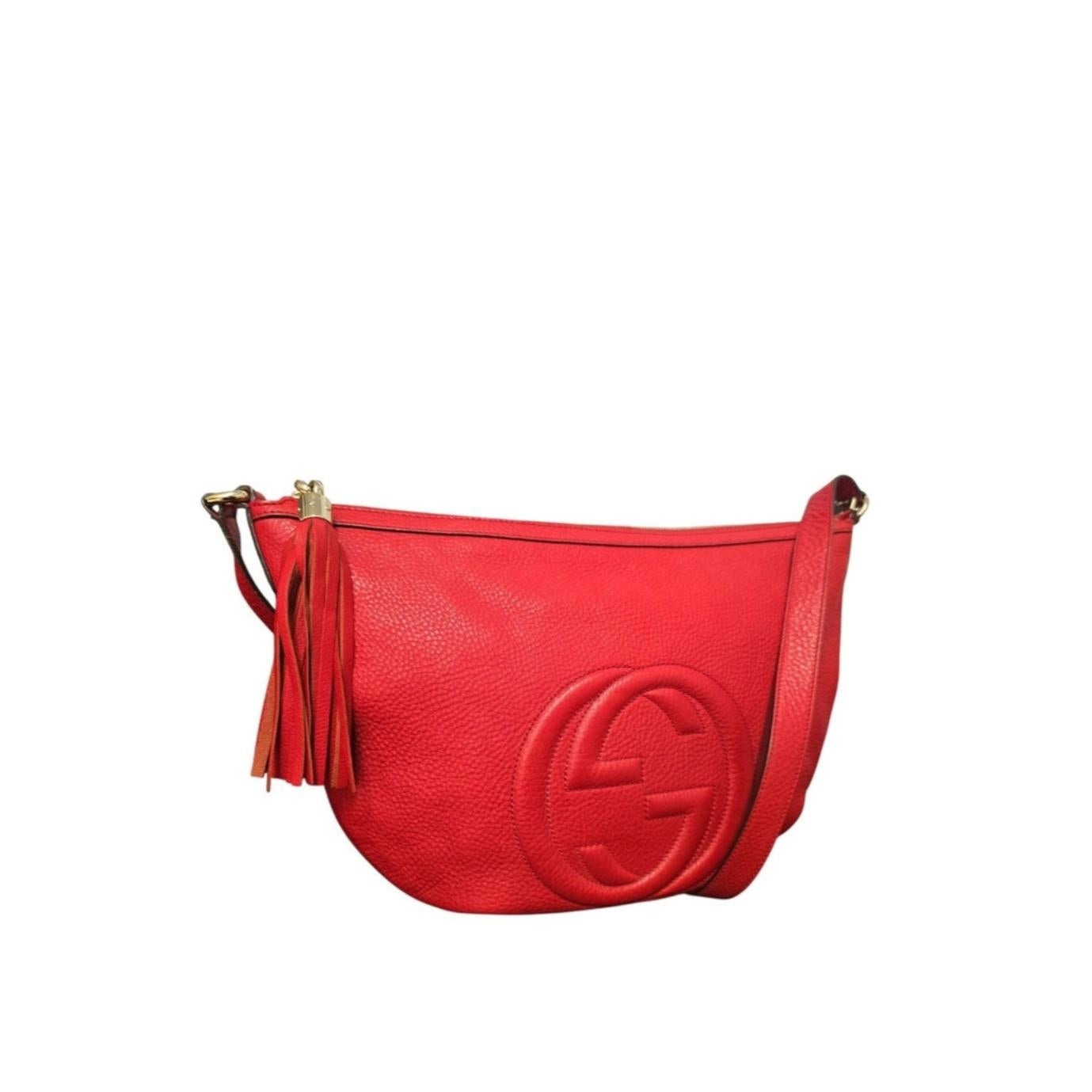 Le sac GG Medium SOHO de Gucci est réalisé dans un luxueux cuir de veau rouge grainé. Il présente une silhouette hobo polyvalente avec une fermeture à glissière simple, rehaussée d'un pompon rouge. Sa bandoulière réglable permet de personnaliser le