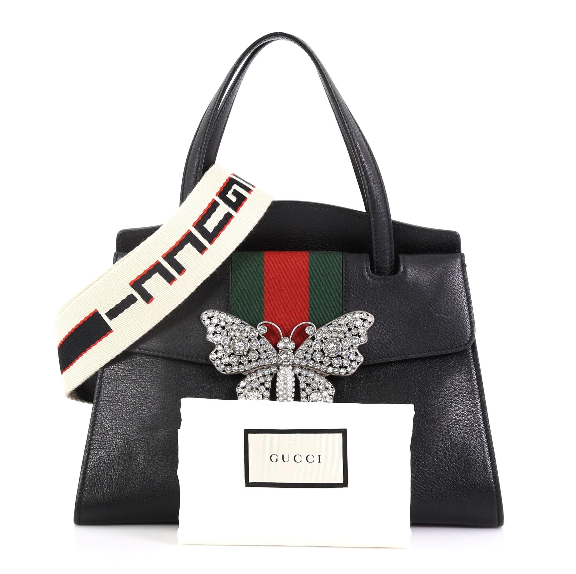 This Gucci Totem Top Handle Bag Leather Medium, crafted from black leather, features dual leather top handles, green and red web with crystal butterfly ornament, and gold-tone hardware. Its press-lock closure opens to a beige microfiber interior