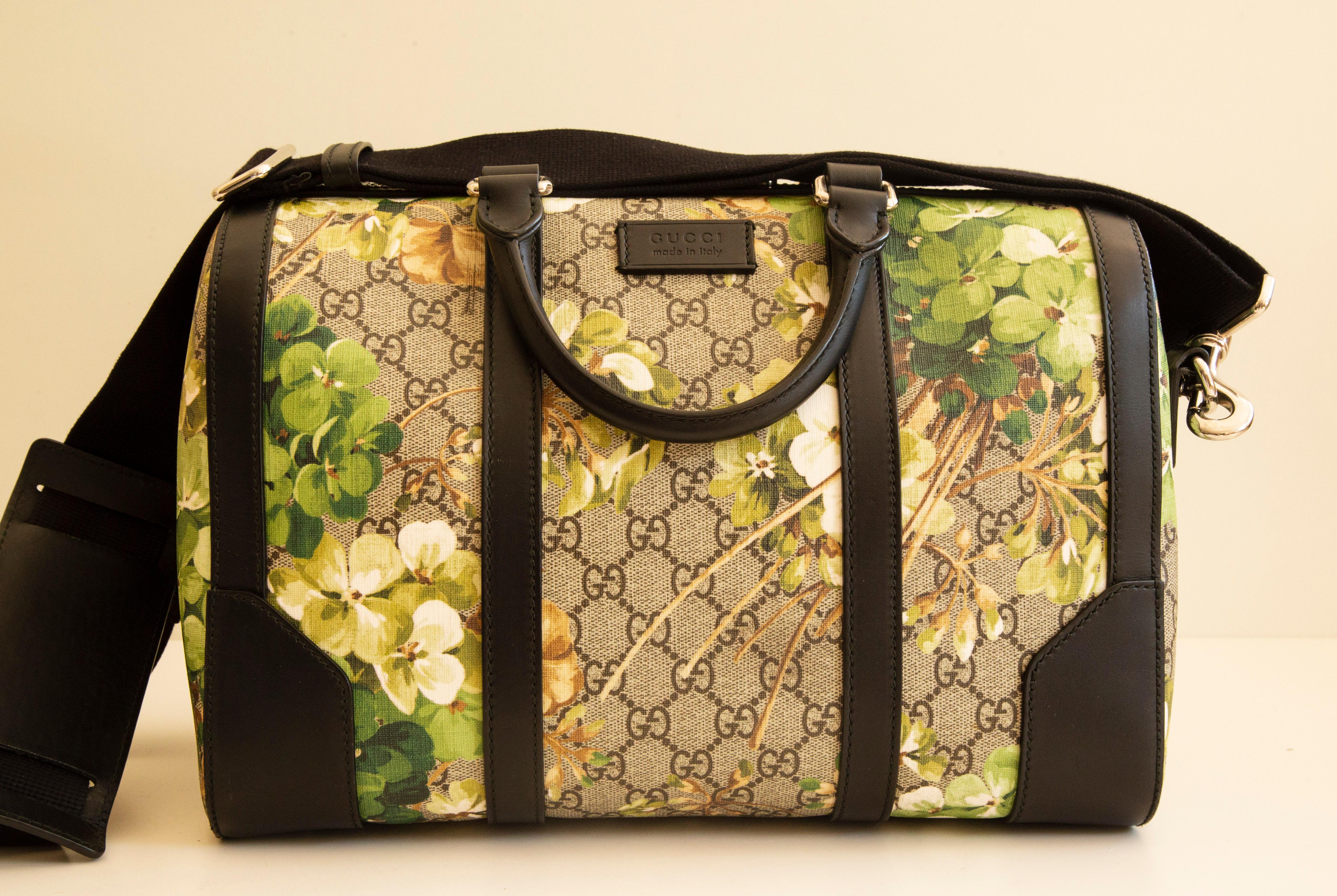 A Gucci duffle convertible handbag/cross body bag with brown green print blooms coated canvas and black leather trim & silver tone hardware. The interior is lined with dark brown synthetic fabric, and next to the major compartment it features two