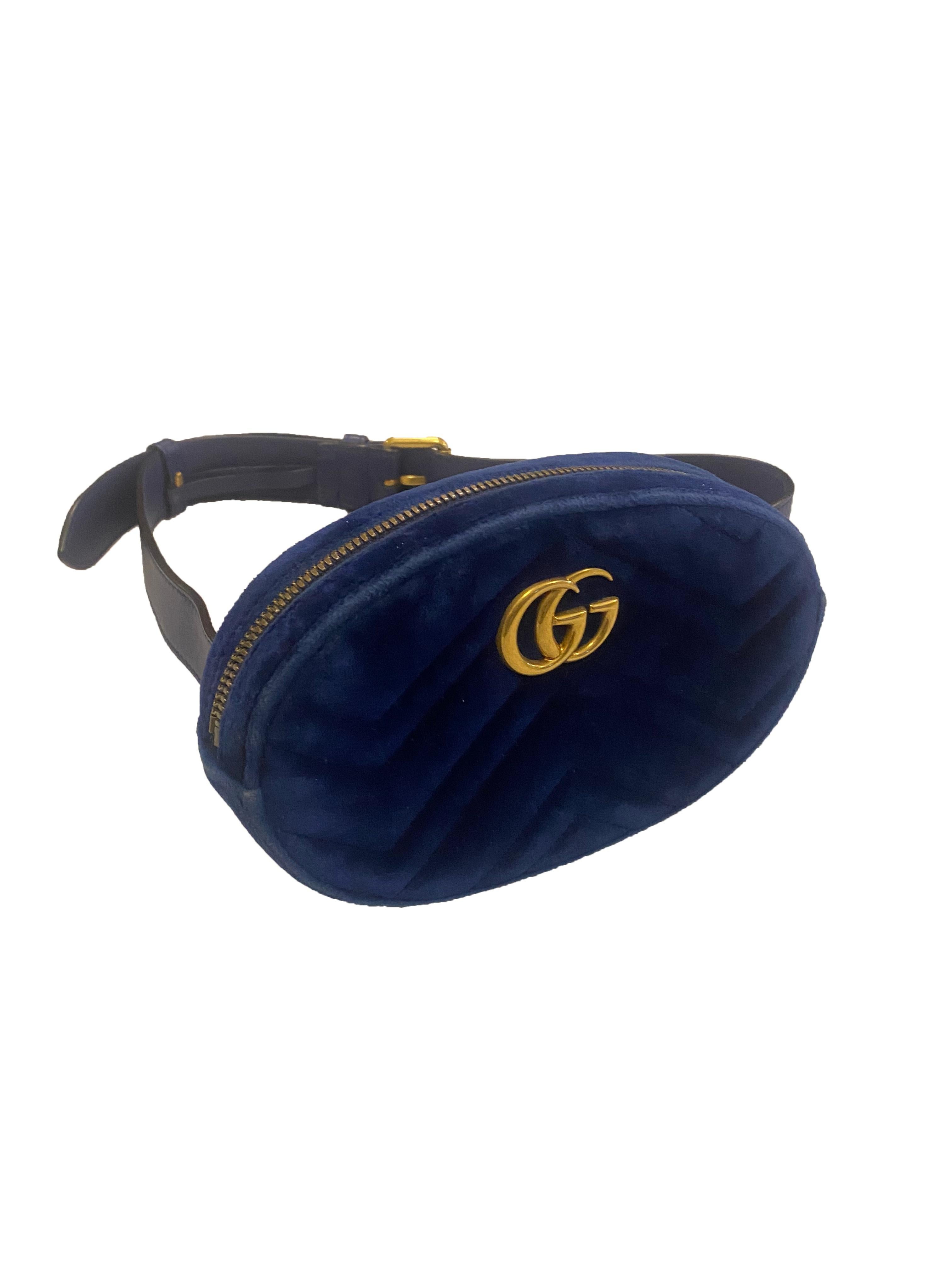 Velvet belt bag from Gucci. Structured, oval, midnight blue velvet pouch with blue leather belt and gold toned hardware. Branded 'GG' on the front in gold metal lettering. Smooth leather belt has 9 buckle settings for adjustable wear. Made in Italy.