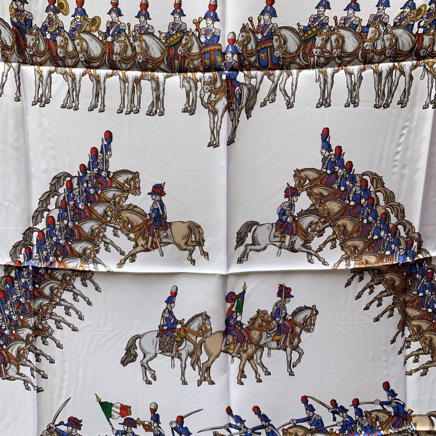 Foulard en soie Vintage Gucci des années 80. Au centre, un imprimé représente un défilé de carabiniers à cheval ; aux coins, on trouve les hatson typiques des carabiniers. Fond blanc et bordures rouges et bleues. 100% soie. Dimensions : 35 x 35