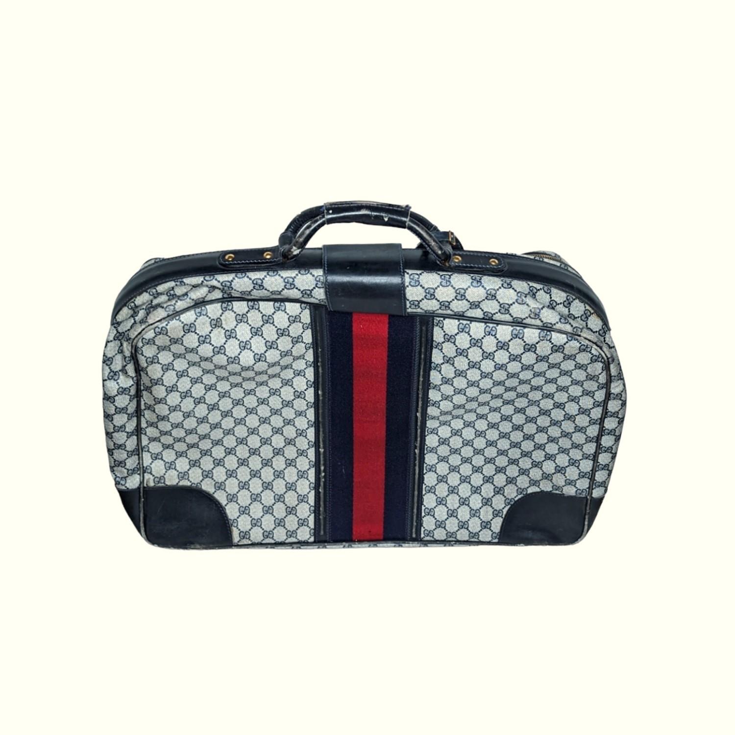 Vintage blue 'GG' monogram coated canvas suitcase from Gucci Circa late 1970’s - early 1980’s. Navy leather trim. Gold-tone hardware details. Navy and red striped textile down the front and back. Two wraparound zippers for closure. Two handles with