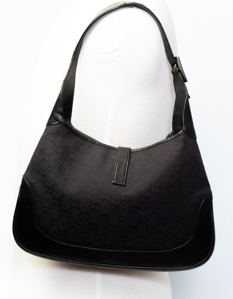 This hobo bag is made of Gucci GG monogram canvas in black with a looping leather shoulder strap, leather base and leather trim. A cross over strap with a squared silver tone piston lock secures the top of the bag. This opens to a black ja interior