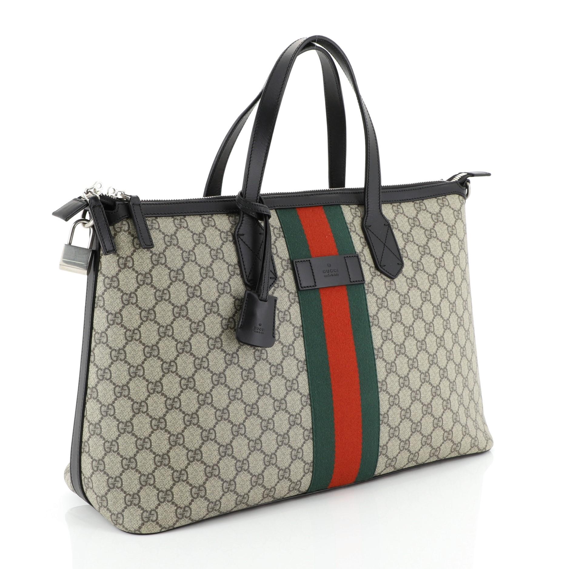 This Gucci Web Duffle Bag GG Coated Canvas Medium, crafted in brown GG coated canvas, features dual flat handles, web strap detailing, leather trim, and aged silver-tone hardware. Its zip closure opens to a black nylon interior with zip