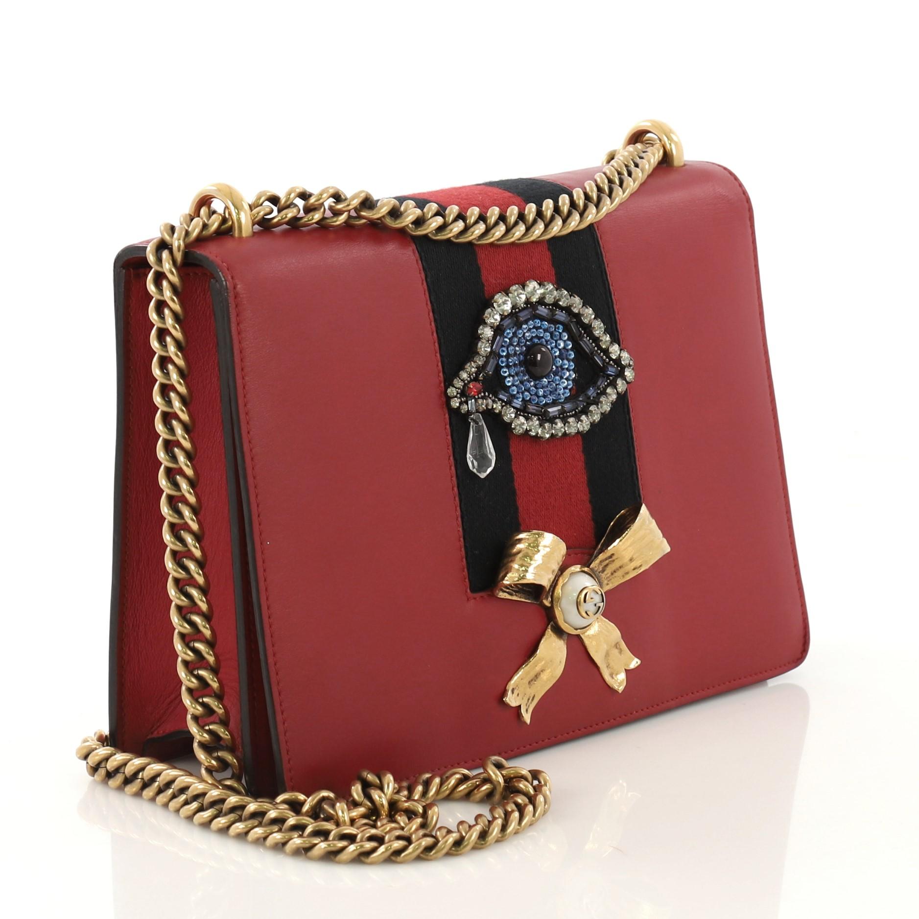 This Gucci Web Peony Chain Shoulder Bag Embellished Leather Medium, crafted in red embellished leather with beaded eye appliqu̩e and golden metal bow with faux pearl, features chain-link shoulder strap, black and red wed stripe and aged gold-tone