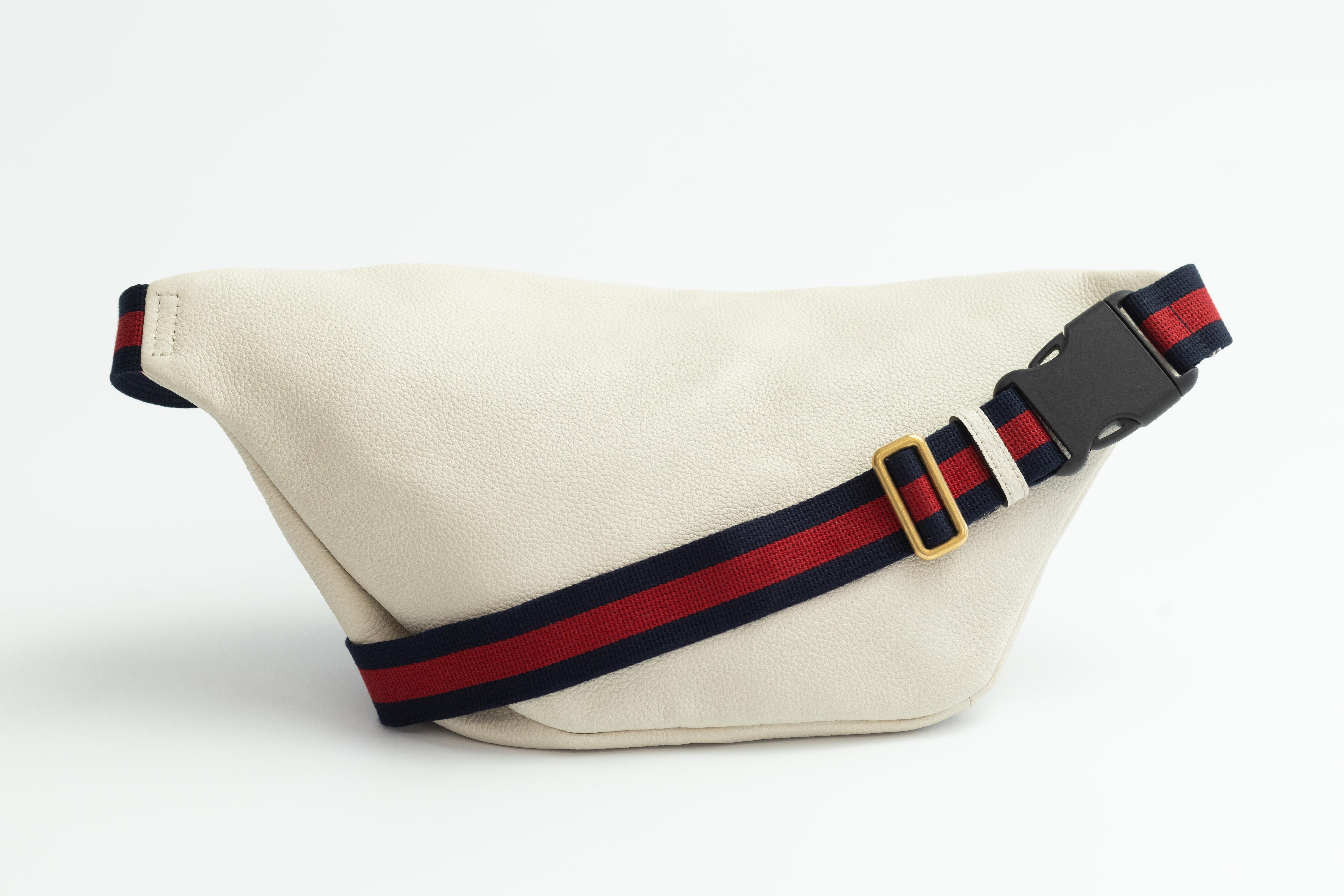 This belt bag is white leather with a vintage Gucci logo. This bag has a nylon navy and red adjustable web belt that can be worn as a belt bag on the hip or waist. The front zipper opens to a beige natural fiber interior.

Color: Off white
Material: