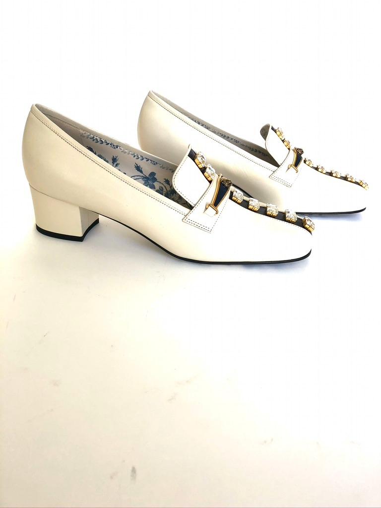 Squared heel loafer, the front of the shoe is enriched with a contrast inlaid stripe dotted with large crystals. An enameled metal detail displays the feline head—one of the most recent House emblems. Ivory leather with dark blue stripe. Blue