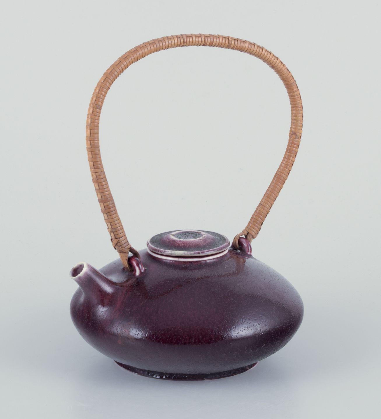 Gudrun Meedom for Bing and Grondahl.
Stoneware teapot decorated with oxblood glaze. 
Bamboo handle wrapped with wicker. Japanese style.
Dating: 1960s
Made and marked at Bing & Grondahl. 
In excellent condition, with bubbles in the glaze from