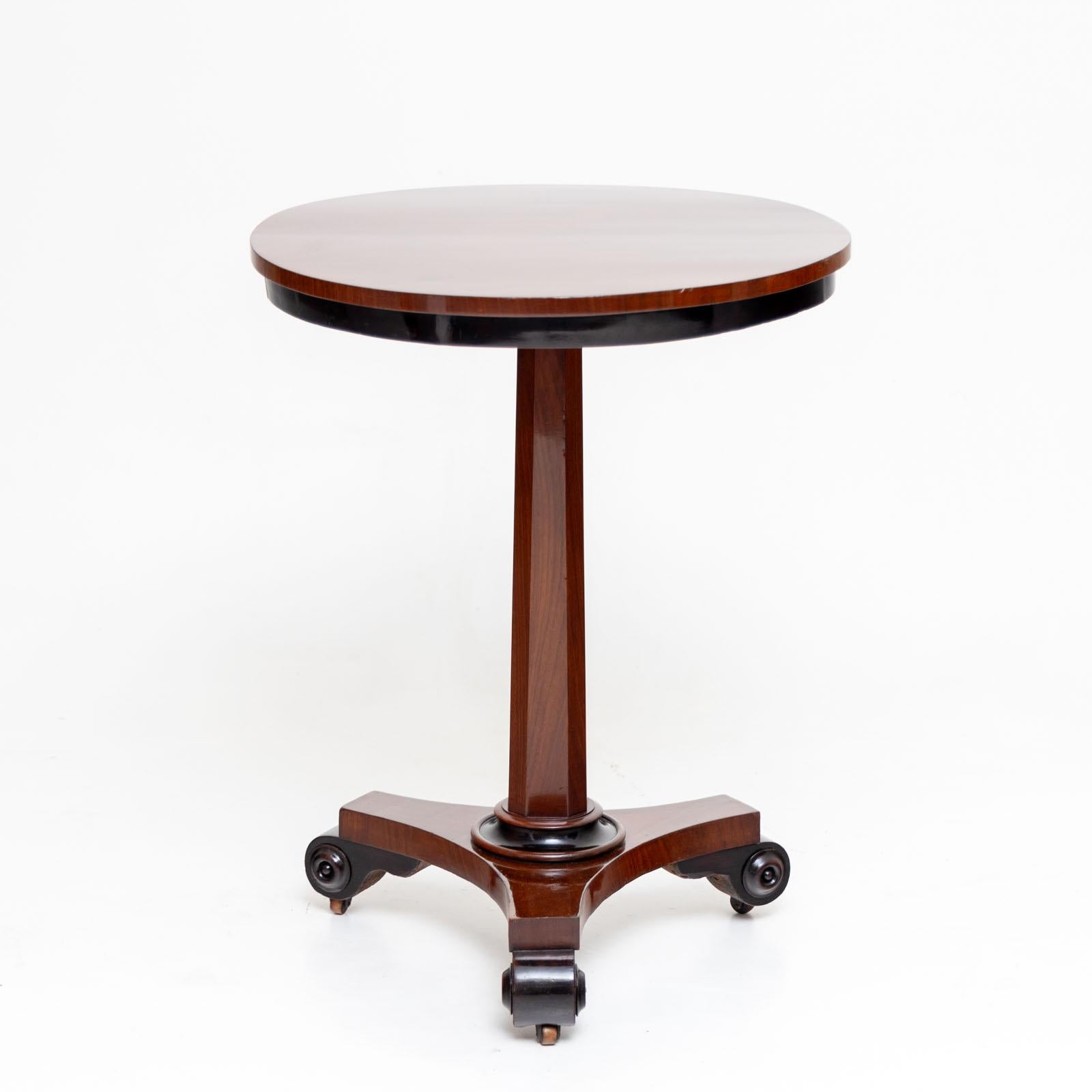 Small gueridon on castors with blackened volute legs and a tripod base. An octagonal, smooth column rises above it, supporting the round table top with a dark frame. The table has been restored and polished by hand.