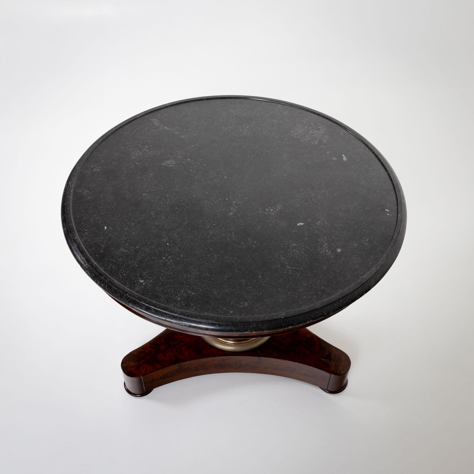Gueridon with round table top made of gray stone and mahogany veneered, conical central column and trefoil base. A twisted brass ring surrounds the central column as a base. The table has been restored and polished by hand. 