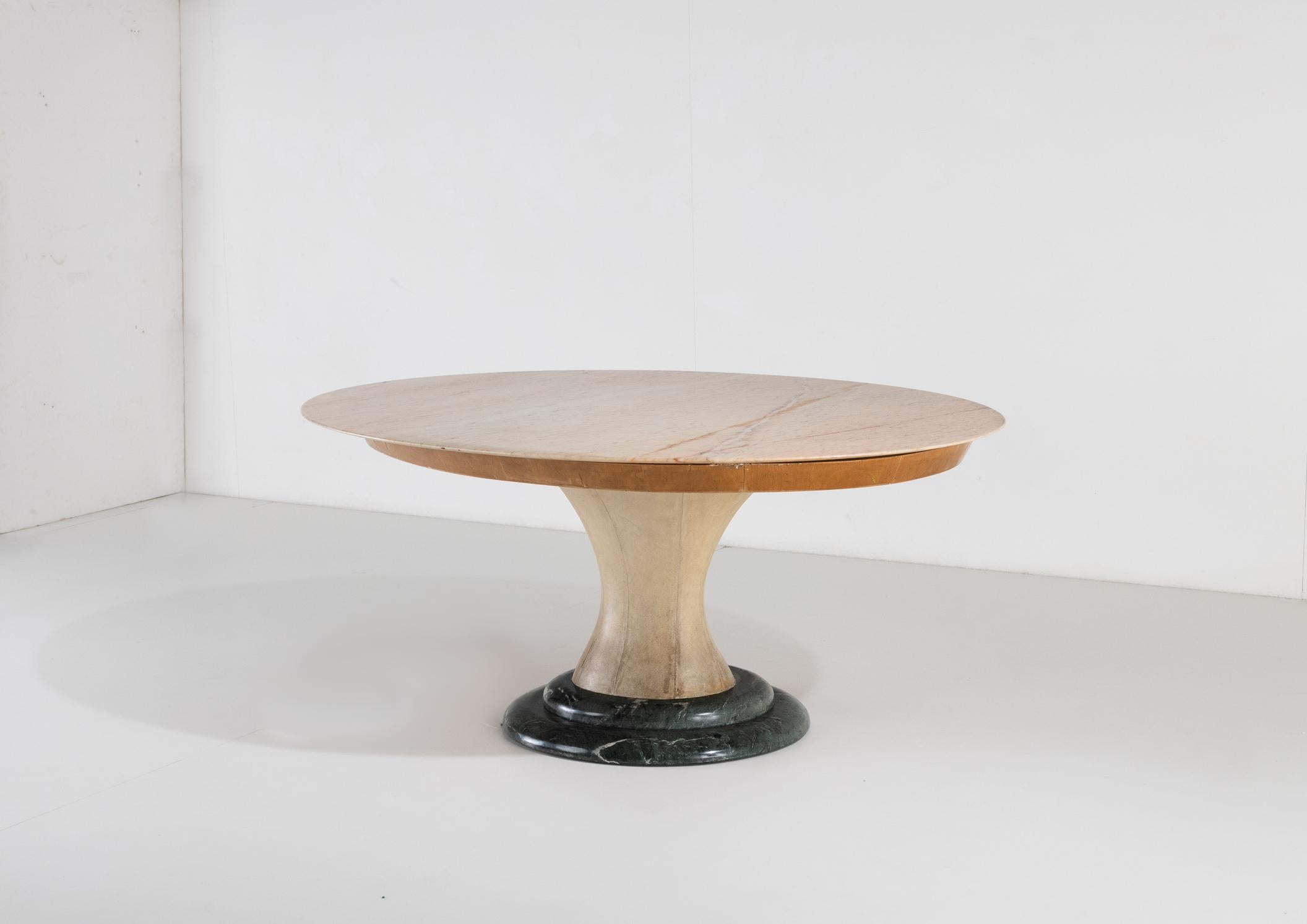 A table with soft and elegant lines, it features a solid base anchored with green marble and an hourglass-shaped wood main body with original parchment covers. The support holds a thick-edged wood top that, for its part, holds a splendid pink oval