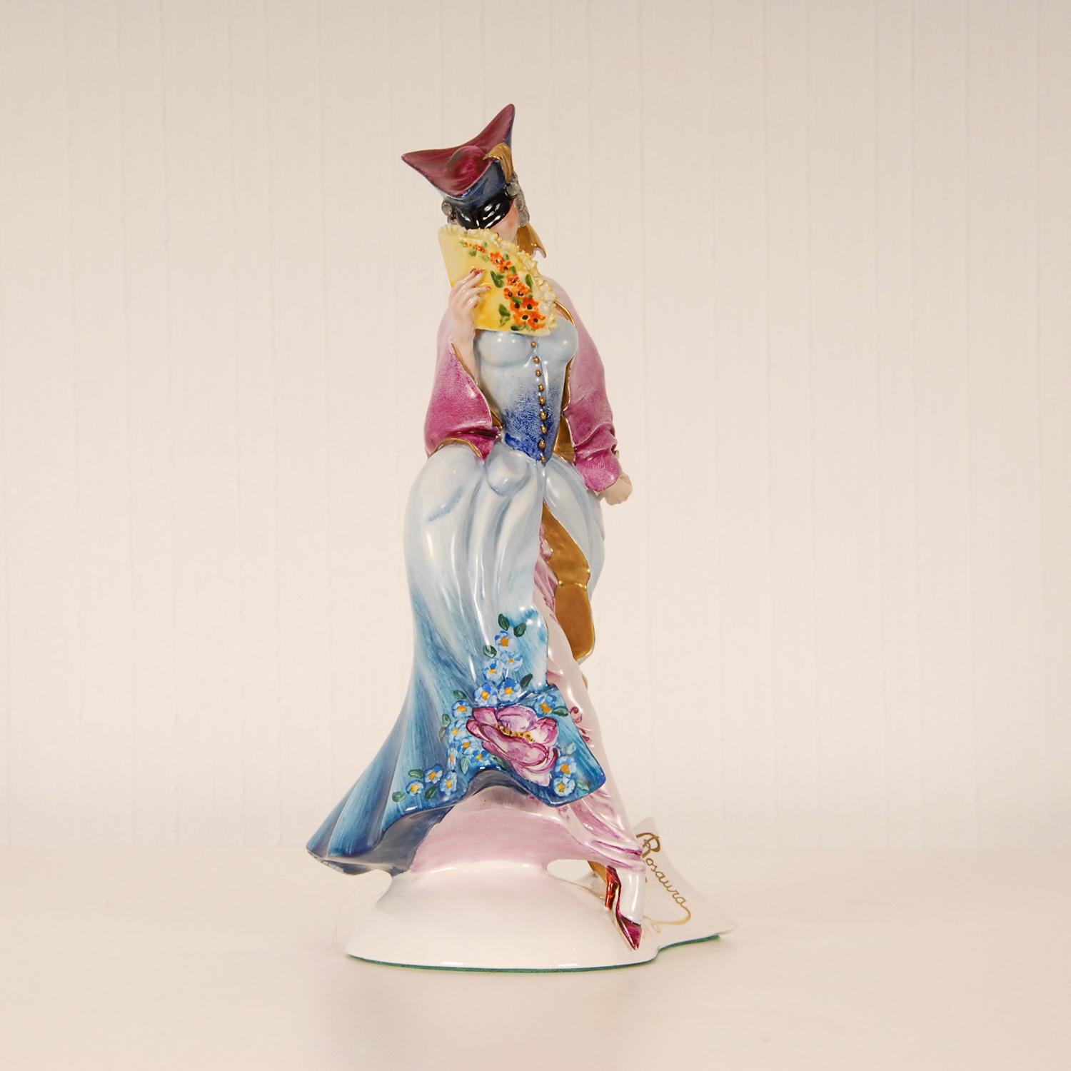 Italian Art deco porcelain figurine
Depicting a Commedia Dell'arte figure Venice Carnaval, with a mask
Hand-crafted figurine her pink dress is heighten with gold and embellished with hand painted flowers 
in bright colours, In her hand a fan
Style
