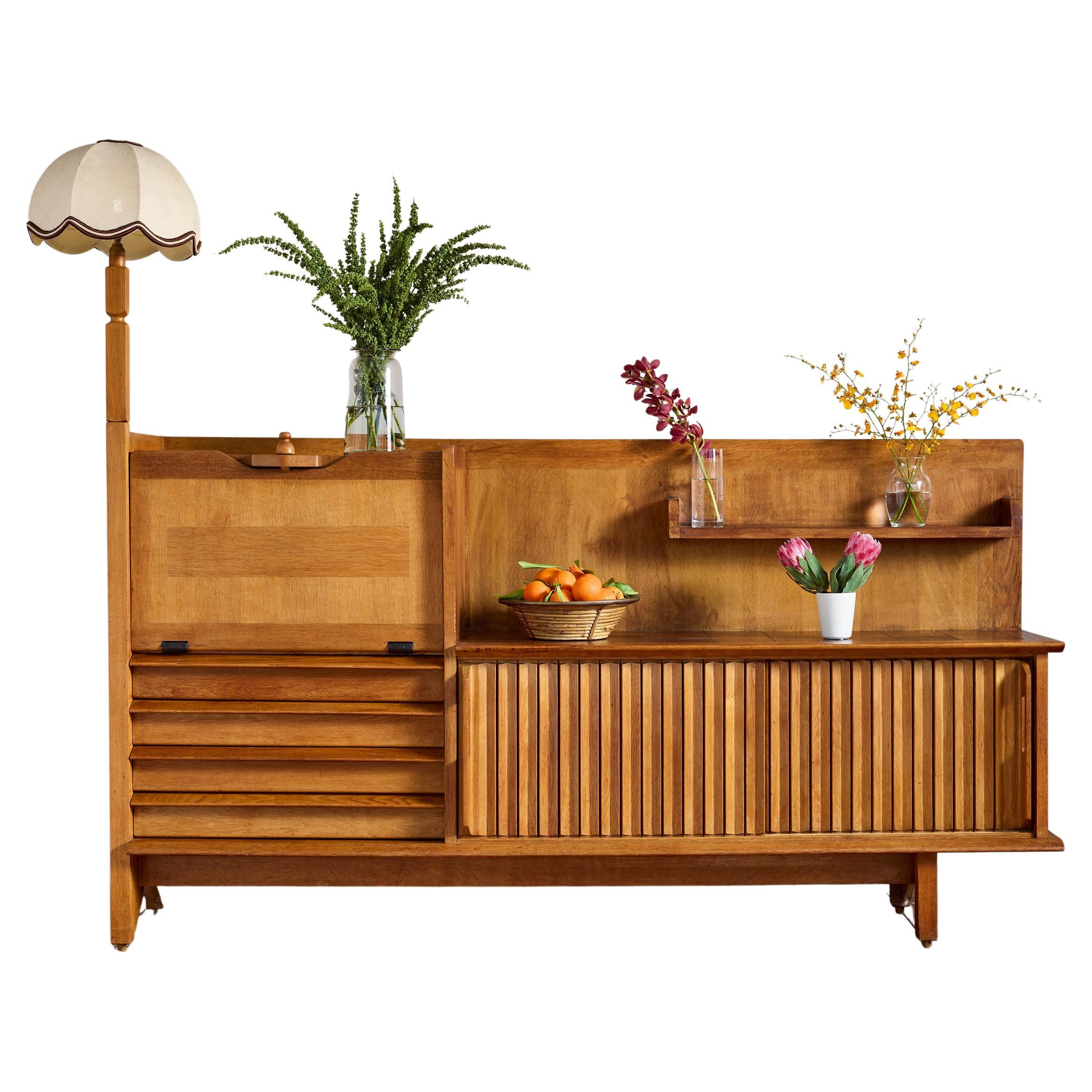 Beautiful sideboard or buffet in solid oak designed by French designer Guillerme et Chambron - France, 1950s

Cabinet has two sliding front doors and four drawers along with a drop down door that reveals open shelving. 

Rare original lamp with