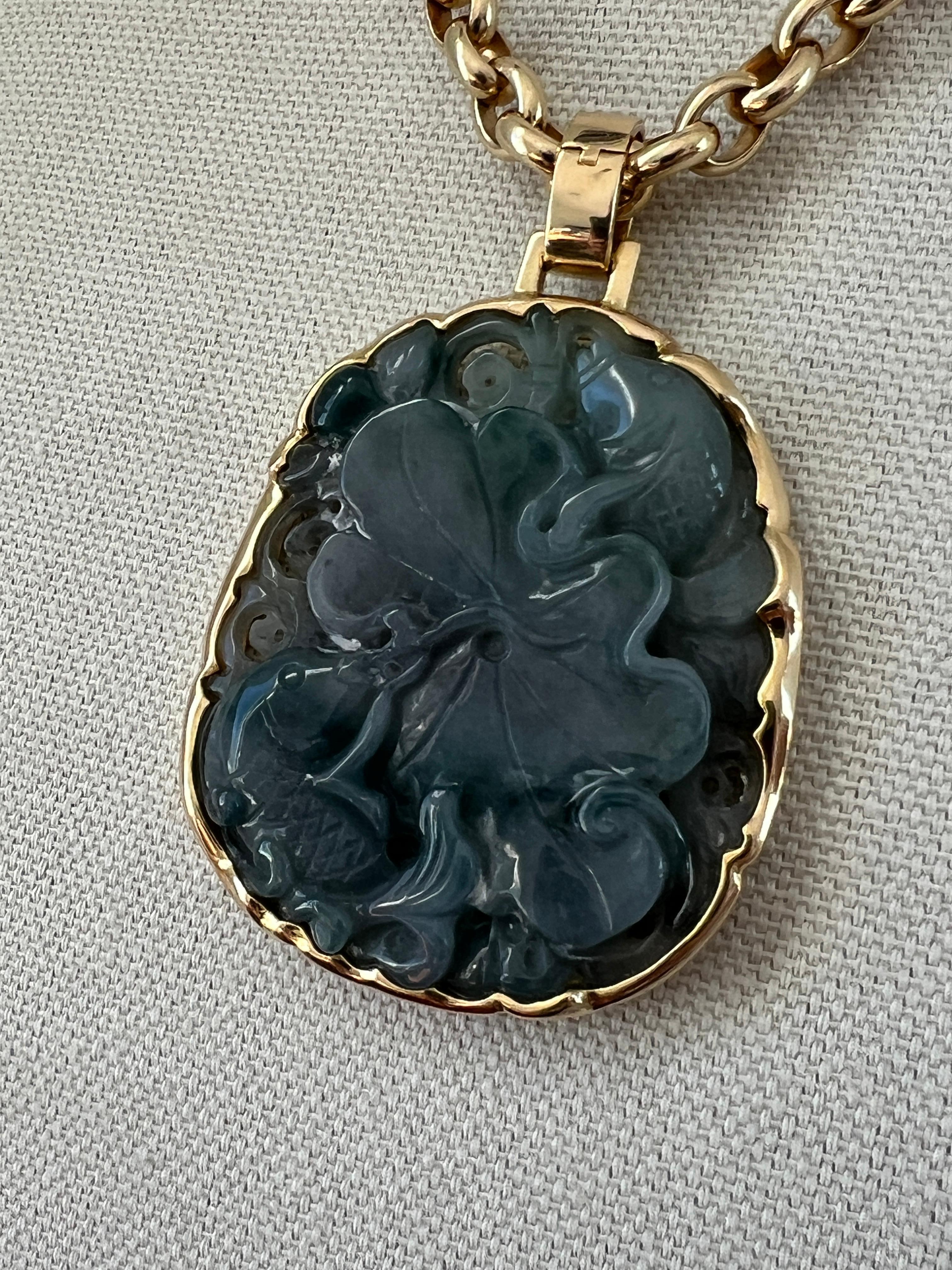 This contemporary Gump's San Francisco pendant features a carved jade centerpiece depicting two fish with an intricate floral design, encased in a 14k yellow gold freeform frame. This elegant piece weighs 51.01 grams measures 2 inches by 1.5 inches