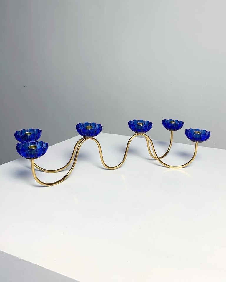 Six armed candelabra designed by Gunnar Ander for Ystad-Metall in Sweden, 1950s.

Made of solid brass with blue pressed glass flowers, marked „made in Sweden“.

Measures: width: 40.5 cm
Depth: 15.5 cm
Height: 10.5 cm
For candles with a