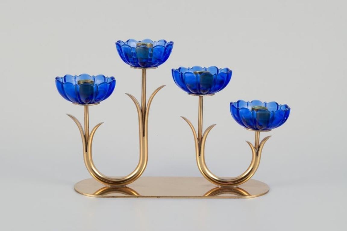 Gunnar Ander for Ystad Metall, Sweden. 
Candlestick holder in brass and blue art glass shaped like flowers. 
For four candles.
From the 1950s.
In excellent condition with good patina.
Marked.
Dimensions: W 20.0 cm x H 13.5 cm.
