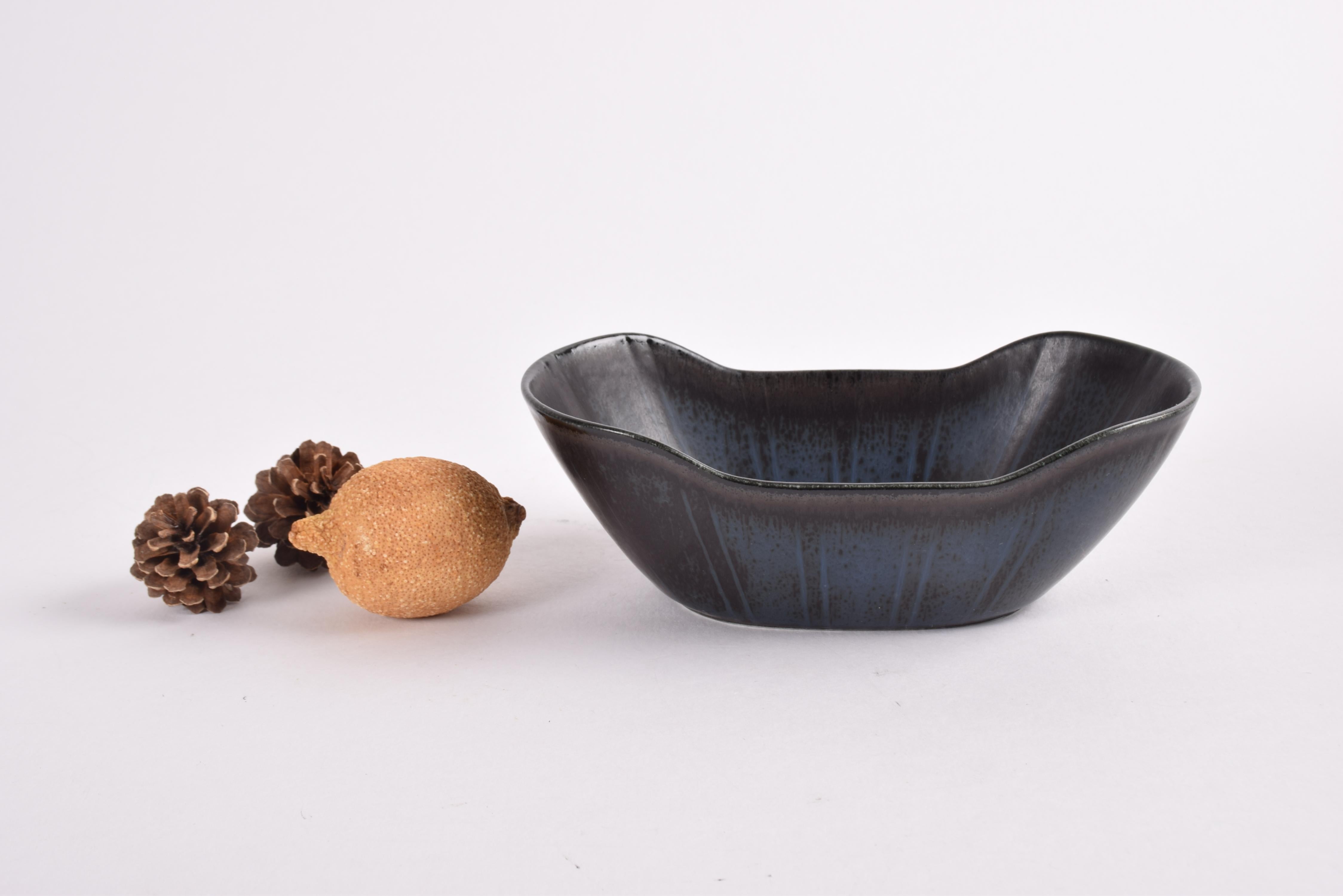 Small oblong ceramic bowl by Gunnar Nylund for Rörstrand Sweden.
Made ca 1950s to 1960s.

The bowl features a glaze with a mix of dark colors: black, brown and blue gray, It is decorated with a stripe decor on the walls and circles on the inside