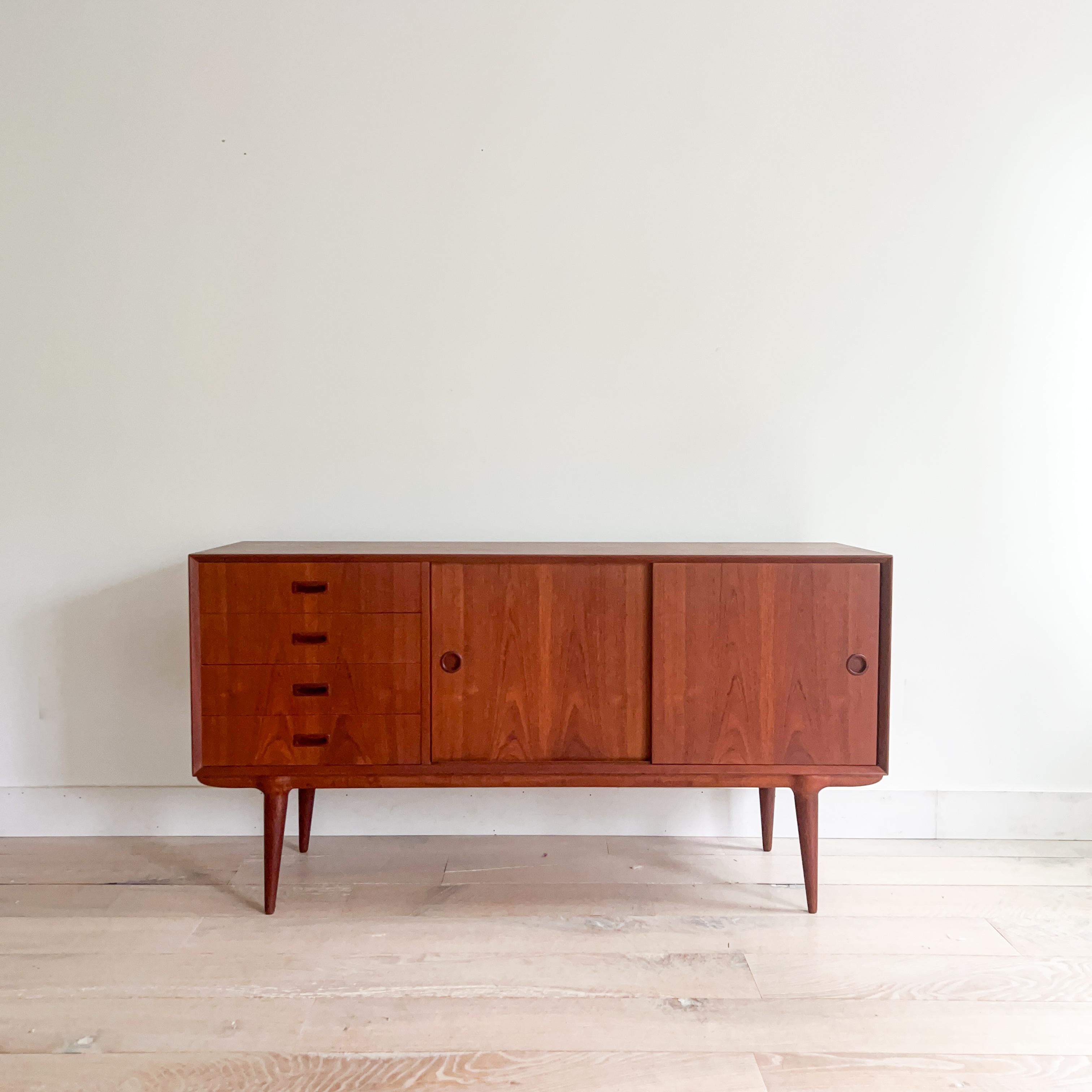 This stunning mid-century modern credenza, designed by Gunni Omann, features elegant teak construction with smooth sliding doors. Adjustable shelving behind the sliding doors offers versatile storage, and the drawers open and close effortlessly.