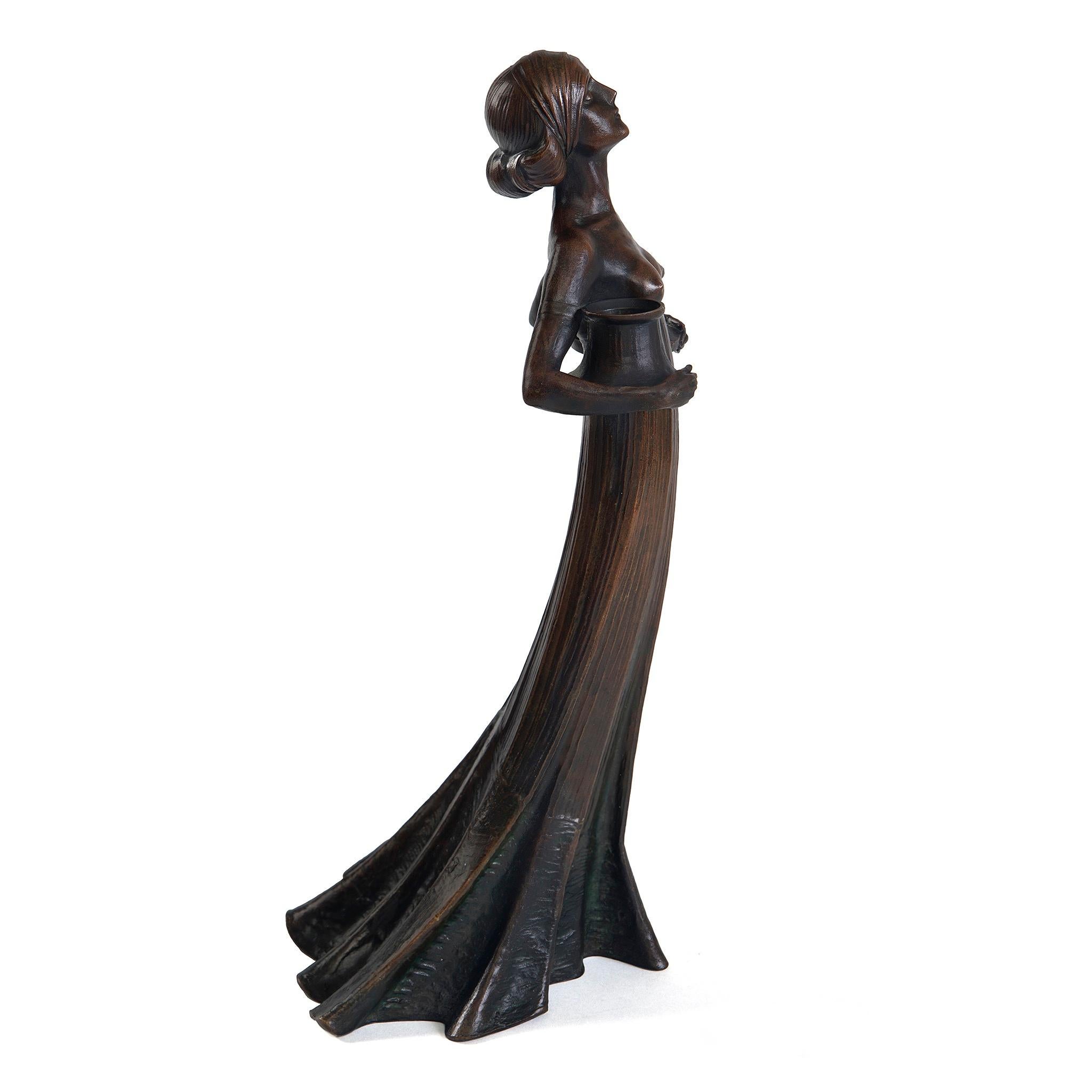 A bronze Art Nouveau figural candelabrum by Gustav Gurschner. A young woman with twin buns, dressed in a long flowing skirt, with her breasts exposed, holds a seed capsule in each arm, into which a candle can be inserted. Gurschner displayed his