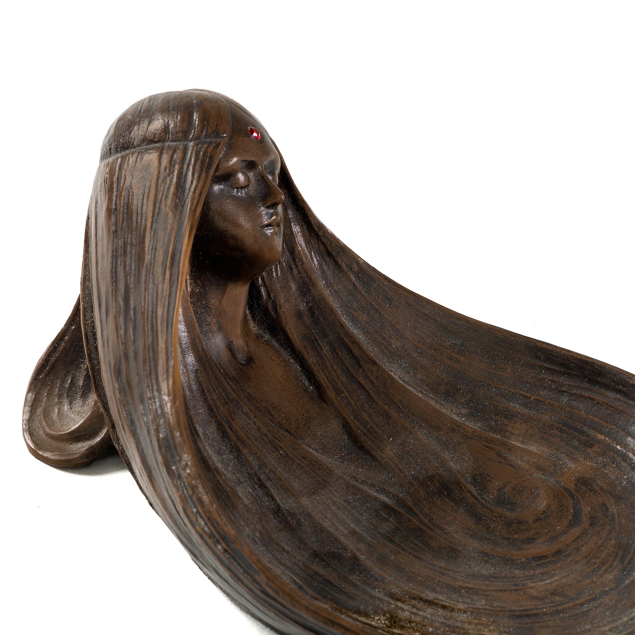 This French Art Nouveau bronze pin tray by Gustav Gurschner titled “Symbolist Reverie” depicts the face of a sleeping maiden wearing a diadem. Her long, flowing hair swirls before her relaxed, sleep-like visage to create the tray. The headpiece she