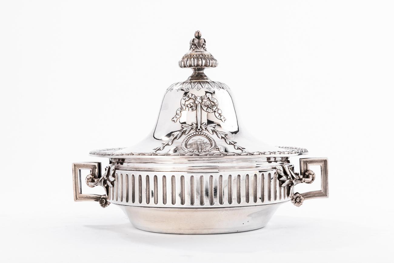Vegetable dish in round shape, in solid silver, on a flat background, the body decorated with fluting is flanked by two geometric handles, the chimney cover is engraved with two beaded and monogrammed medallions, the socket is placed on a terrace of