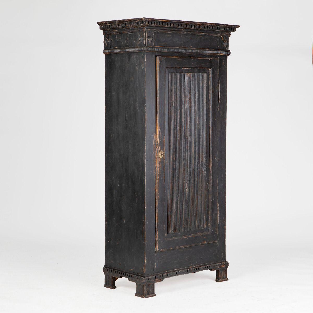 Armadio Gustavo a un'anta in pino verniciato nero. Il mobile è decorato con i classici dettagli intagliati a mano tipici dell'epoca, come l'intaglio a dentello sotto la modanatura superiore e il grembiule, gli ornamenti a piramide e i pannelli