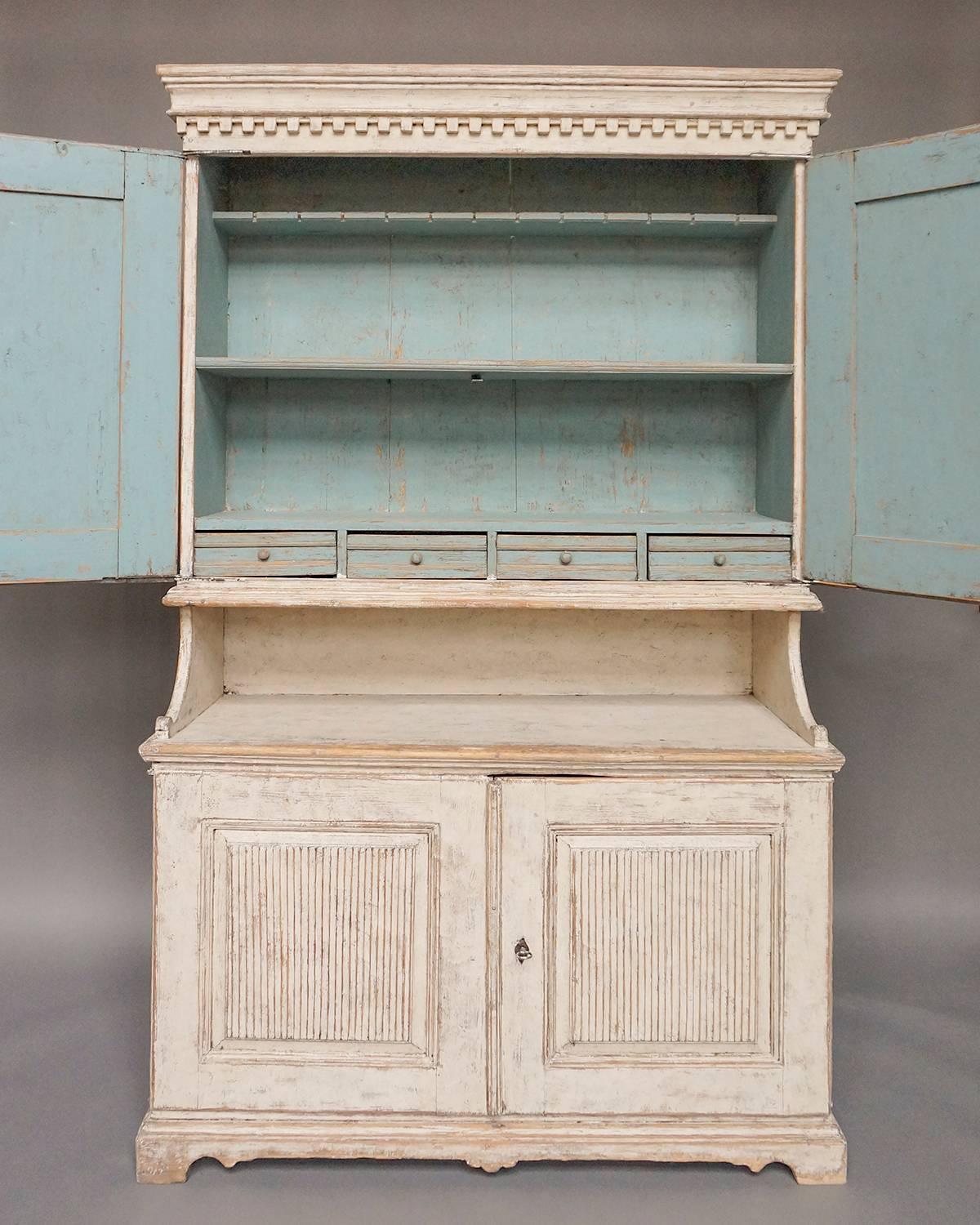 Period stepback cabinet in two parts from Värmland, Sweden, circa 1820. The upper section has two paneled doors with reeded lozenges under a cornice with dentil molding. The interior has two shelves, one notched for spoons, and four small drawers.