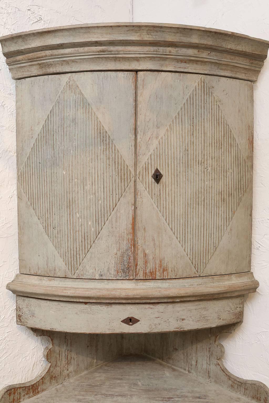 Gustavian corner cabinet from Sweden. The gray-green painted surface of this cupboard bears evidence of hand-planning and 18th century Provincial craftsmanship. It features a carved cornice, middle shelf, upper and lower convex double doors and a