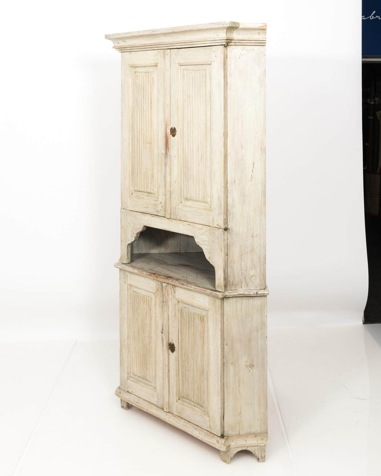 White painted Gustavian style corner cupboard with carved wood doors, ribbed panels, and brass escuctcheon, circa 1890-1920.
    